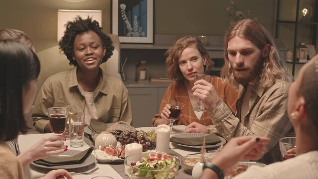 Medium close-up of six young multiethnic friends sitting at dinner table at home, talking and laughing, then raising wine glasses, clinking and smiling