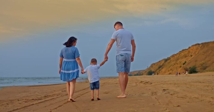 Married couple with a child are walking by the sea, holding the baby by the hands and lifting him up. Parents walk with the baby along the shore, stepping with their bare feet on the sand. Beautiful