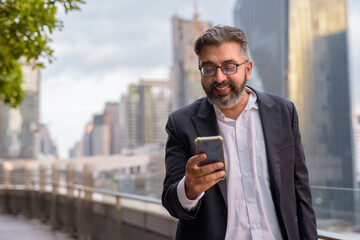 Portrait of handsome Indian businessman in city at rooftop garden using phone