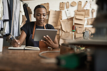 African female leather worker writing notes and using a tablet