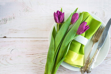 Spring or summer table setting with vintage fork and knife, green napkin with ring, bouquet of purple tulip flowers. Holidays background with copy space