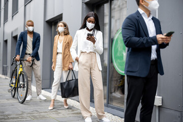 Fototapeta na wymiar Multiracial businesspeople in medical masks waiting in queue on city street. Concept of business during Coronavirus pandemic. Idea of break on job. Businessmen and businesswomen wearing formal wear