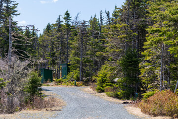 Fototapeta na wymiar Both a male and female green wooden outhouses in the woods off a gravel road in an RV park. The road has forest on both sides. There are campsite markers along the path. The sky is blue with clouds.