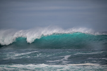 Obraz premium An angry turquoise green color massive rip curl of a wave as it rolls along the ocean. The white mist and froth from the wave are foamy and fluffy. The Atlantic Ocean in the background is deep blue