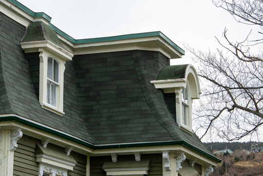 The roof of a vintage green wooden building with cream color trim, multiple double hung windows, dormers, and a green shingled hip roof. It's an old wooden house with multiple stories. 