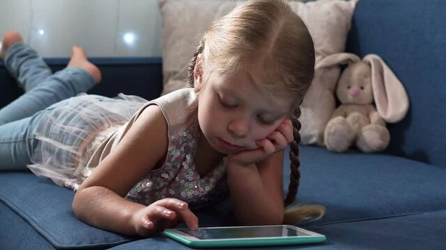 A beautiful blonde girl with two pigtails in a smart jacket and dzheinski lies on a blue sofa and plays with a tablet. The child looks into the gadget. The girl scrolls through the pictures