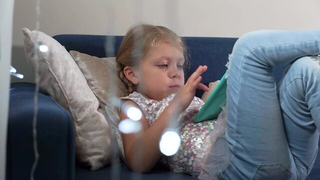 A beautiful blonde girl with two pigtails in a smart jacket and dzheinski lies on a blue sofa and plays with a tablet. The child looks into the gadget. The girl scrolls through the pictures