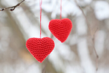 Fototapeta premium Love hearts hanging on a tree branch in winter forest. Two red knitted Valentine hearts, symbol of romantic love, background for holiday