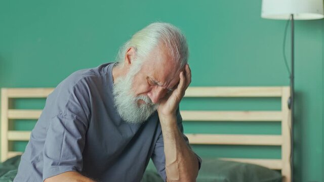 Tired Elderly Man Has Acute Headache While Sitting on ahe Bed At Home. Virus, Headache, Migraine, Anxiety. Seniors, Dizziness, High Blood Pressure, Health Care and Retirement Insurance.