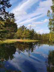 The mirror surface of a forest lake, in which trees and the sky with beautiful clouds are reflected.