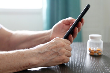 Elderly woman with smartphone and pills sitting at the table, selective focus on wrinkled female hands. Concept of online communication at retirement, search for drug properties, call an ambulance