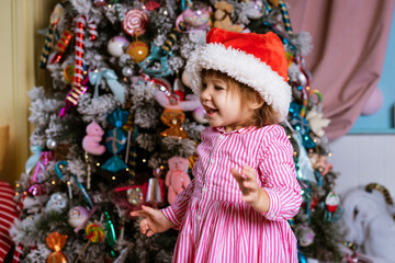Funny child little girl in santa claus hat and pink dress laughs with happiness on bright festive background of decorated christmas tree. Merry christmas and new year holidays concept