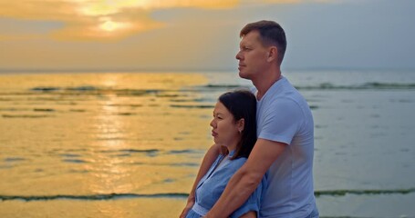Beautiful married couple is standing by the shore and dreaming. Man carefully hugs his pregnant wife, a little son is playing next to her. Family waiting for a baby, happy motherhood and childhood. 4k