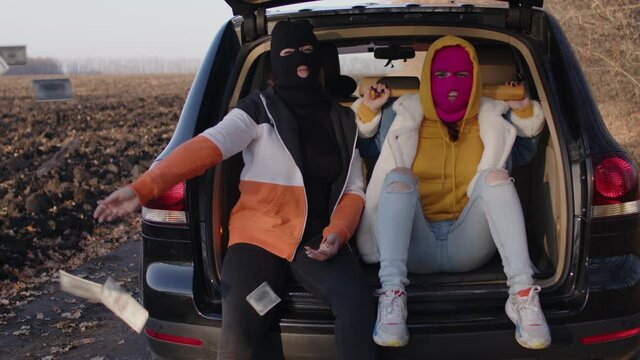 Women in balaclava sitting in trunk of car. Hooligans in masks. One female with baseball bat, other throws dollar bills.