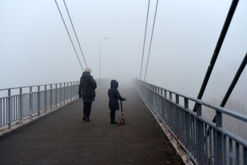 A woman and a girl with a scooter walk forward on a pedestrian bridge. There's a thick fog all...