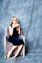 Obraz premium Attractive happy young woman holding a cup of tea or coffee sitting in armchair, on blue fabric background
