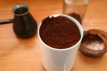 Electric coffee grinder with white plastic case, open transparent lid and crushed ground fresh aromatic roasted coffee beans on brown table top background with turk, glass jar, closeup