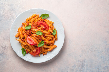 Penne pasta with bolognese sauce, fresh tomatoes and basil in a white plate on a light pink background.