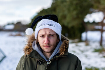 A grumpy young man wearing a black santa hat with the words Bah Humbug written on it