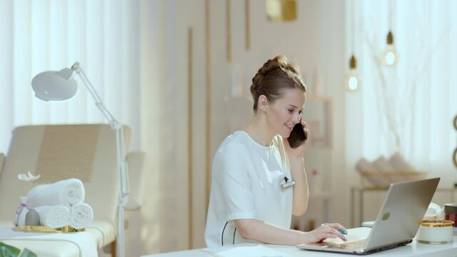 happy woman worker with laptop speaking on a smartphone in modern beauty salon.