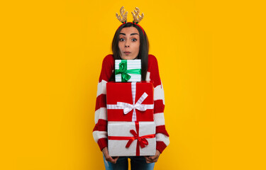 Happy excited young brunette woman in deer hat, with many colorful Christmas gift boxes in hands...