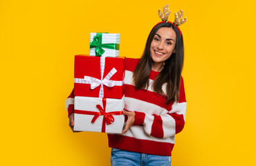 Happy excited young brunette woman in deer hat, with many colorful Christmas gift boxes in hands isolated on yellow background.