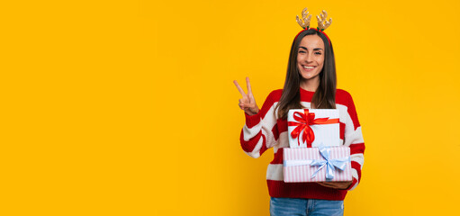 Cute smiling excited young brunette woman in deer hat, with many colorful Christmas gift boxes in hand is showing victory sign isolated on yellow background.