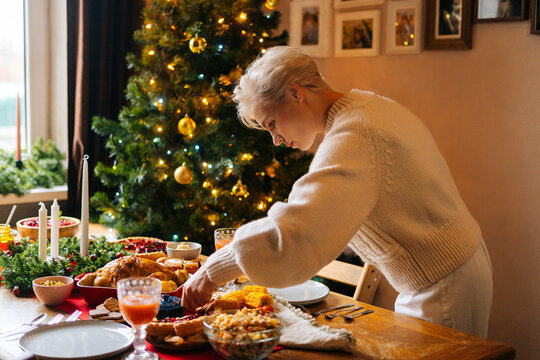 Happy Young Woman Preparing Christmas Dinner Table At Home For Family Party On Blurred Background Of Decorating Xmas Tree And Celebration Lights. Pretty Female Serving Tableware Waiting For Friends.