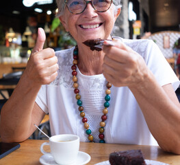 Portrait of senior smiling woman having break at coffee shop with chocolate cake and coffee cup. Carefree elderly woman with eyeglasses and necklace. Thumb up as positive moment - obrazy, fototapety, plakaty