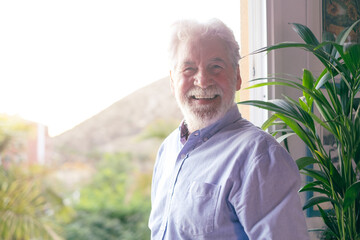 Senior bearded man standing next to the window looking at camera. Caucasian white haired elderly people