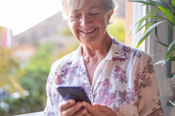 Attractive senior woman at the window holding mobile phone while writing a message. Smiling gray-haired senior enjoying technology and social