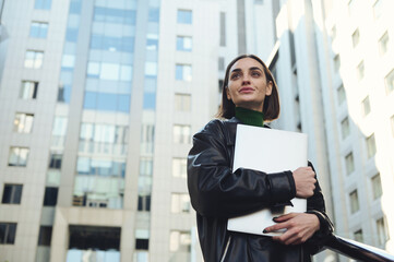 Lifestyle portrait of successful self-confident motivated young business person, attractive freelance woman holding laptop, looking up posing against modern corporate high-rise buildings background © Taras Grebinets