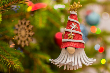 A Christmas tree toy in the form of a New Year's gnome hangs on the tree against the background of garlands.Good New Year spirit.Layout for postcards,place for text,congratulations,invitations.