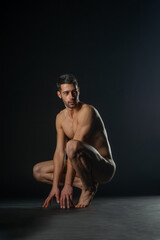 Portrait of a naked male model posing squatted on black studio background