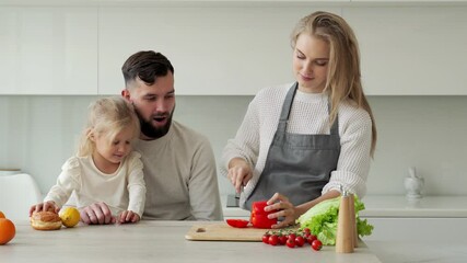 Young family cooks food in the kitchen at home. Portrait of a smiling mother slicing pepper with a knife, next to a bearded father and a little daughter