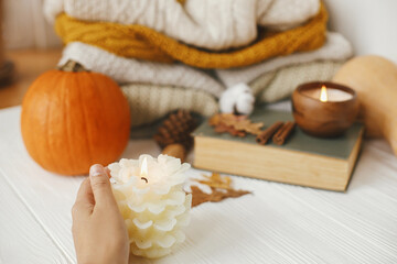 Hand holding burning candle on background of pumpkin, cozy sweaters, autumn leaves and vintage book on white wooden background in room. Hello autumn, cozy slow living. Happy Thanksgiving