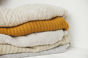 Pile of white, yellow and beige knit sweaters close up in white scandinavian room. Cozy knitted sweaters. Hello autumn. Stylish wardrobe for cold season. Space for text