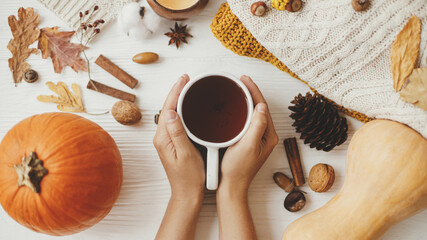 Hello autumn, cozy slow living. Hands holding warm cup of tea on background of autumn leaves, pumpkin, cozy sweaters, burning candle on white wood. Happy Thanksgiving. Flat lay