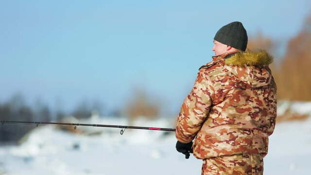An athlete angler spins a spinning reel on a winter fishing trip. He is dressed in a special winter suit and a knitted hat. Sunny frosty day outside the city.