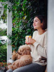 Young asian woman resting outdoors with cup of coffee on porch of country house, chilling outside with poodle dog . autumn lifestyle, leisure free time concept. Copy space