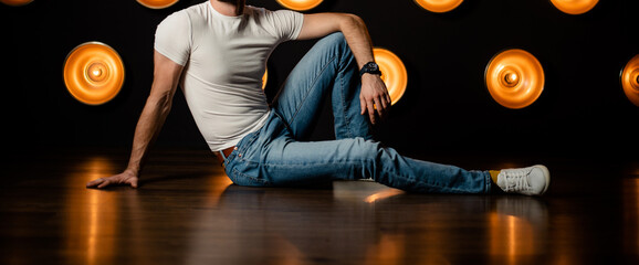 Stylish fashionable unrecognizable man in jeans and a white T-shirt sits on the floor in a dark...