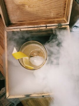 Overhead View Of A Whisky Cocktail With Ice Cube Sphere And Lemon In A Box Filled With Smoke