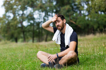 Naklejka na ściany i meble Exhausted after workout training or jogging athlete man sitting outdoors with a towel. Portrait sweaty fitness male in city park background forest and trees. Caucasian sportsman resting after running