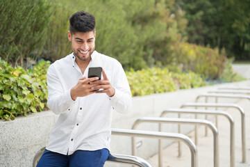 Obraz premium Handsome mixed race young man, smiling, using a mobile phone outdoors. High quality photo