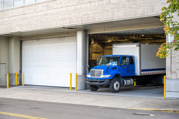 Blue day cab rig semi truck with box trailer unloading cargo standing in dock inside multilevel...