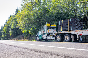 Classic American big rig bonnet semi truck transporting cargo on flat bed semi trailer running on the road with forest on the side