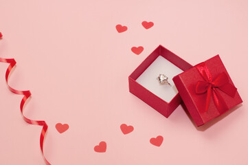 red box with a white gold ring with a precious stone on a pink background with confetti of hearts and a red ribbon