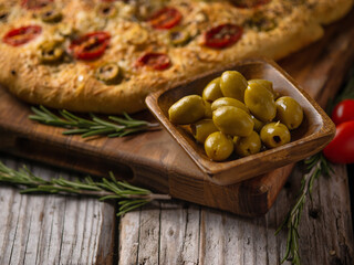 Beautiful composition, close-up. Italian traditional focaccia bread on a cutting board, a bowl of green olives and a sprig of rosemary. Wooden texture. Country style. Instagram, culinary blog.