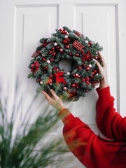 Christmas wreath with red balls and toys in female hands. Frontal view on a white wooden door. New year and christmas concept