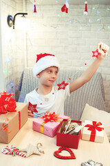 A child in a Santa Claus hat is holding a Christmas tree decoration in the form of snowflakes. Next to the boy are Christmas gifts in boxes. Made by hand. Bright interior. Vertical orientation.